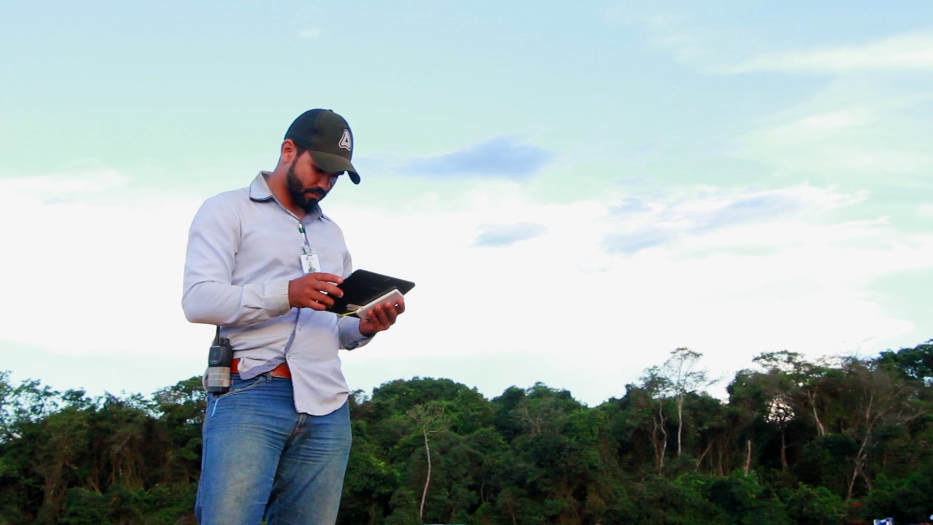 Produtor segurando tablet na mão. No fundo, árvores e um céu limpo e claro