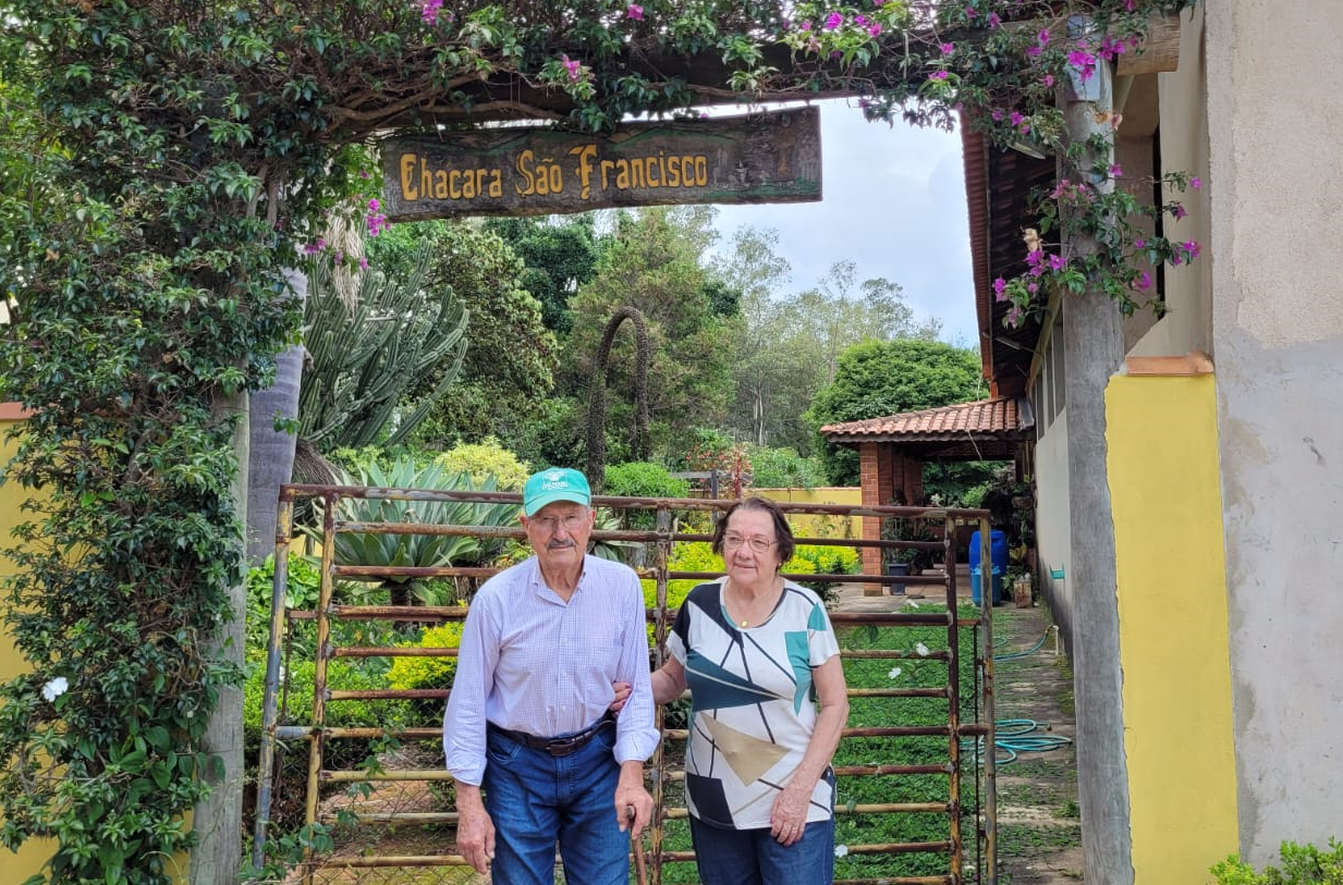 Um casal de idosos em frente a uma porteira de ferro. Sobre a cabeça há uma placa que indica o nome do sítio.