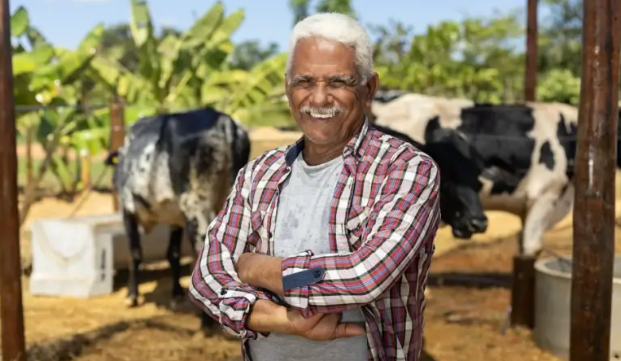 Homem de braços cruzados. No fundo há um pasto com algumas vacas