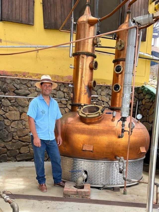 Homem vestido de camisa azul, calça jeans e chapéu. Ao seu lado, há um caldeirão para decantar a cana-de-açúcar