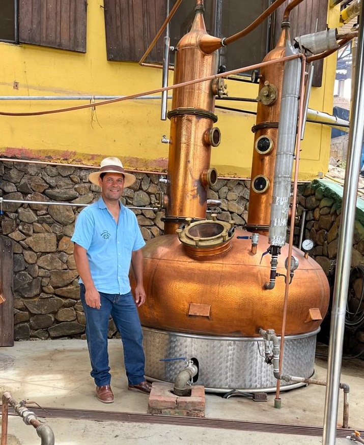 Homem vestido de camisa azul, calça jeans e chapéu. Ao seu lado, há um caldeirão para decantar a cana-de-açúcar