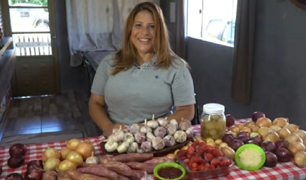 Em uma casa, uma mulher loura e usando uma camiseta cinza. À frente uma mesa com produtor orgânicos que ela produz, em especial alho e cebola