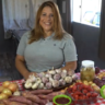 Em uma casa, uma mulher loura e usando uma camiseta cinza. À frente uma mesa com produtor orgânicos que ela produz, em especial alho e cebola