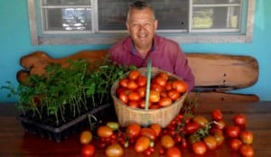 Homem sentado em um banco de madeira. Ao fundo, há uma janela e à sua frente uma mesa replera de tomates