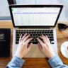 Um notebook e apenas as mãos de um mulher digitando. À direita da foto, há uma xícara com café e óculos escuros. À esquerda, há um caderno de anaotações.