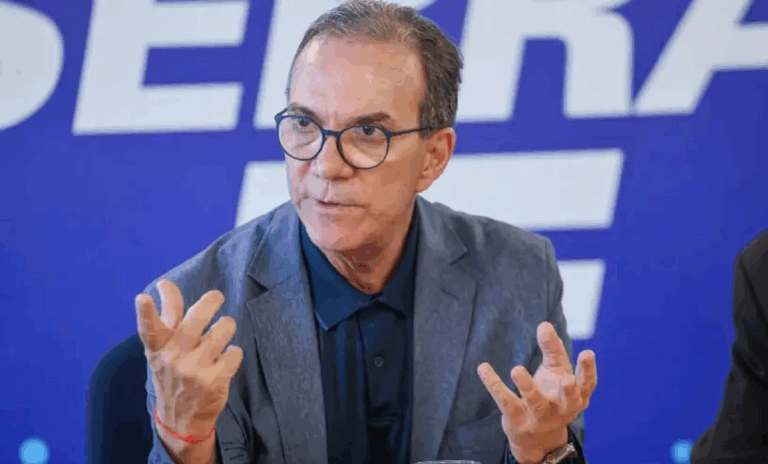 Foto do presidente do Sebrae. É um home que usa óculos e está sentado - gesticulando com as mãos. Ao fundo, há um painel escrito Sebrae.