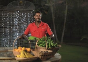 A foto foi tirada à noite. No ângulo central, há um homem com uma blusa vermelha e à sua frente frutas que ele cultiva