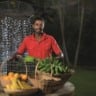 A foto foi tirada à noite. No ângulo central, há um homem com uma blusa vermelha e à sua frente frutas que ele cultiva