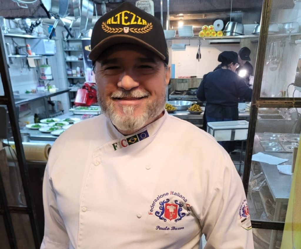 Chef de cozinha. Ele está com um avental branco com o logo do restaurante à esquerda e usa um boné preto.