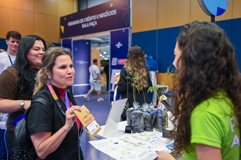 Na foto, há em primeiro plano uma mulher apresentando a credencial para entrar na Feira do Empreendedor 2025, que aconteceu em Curitiba/PR