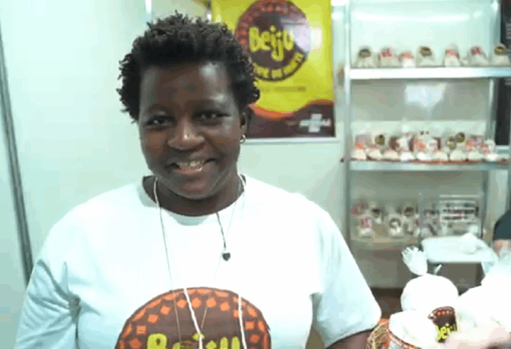 Na foto, aparece uma mulher com um sorriso no rosto. Ela está de camiseta branca e ao fundo há seus produtos artesanais à base de beiju
